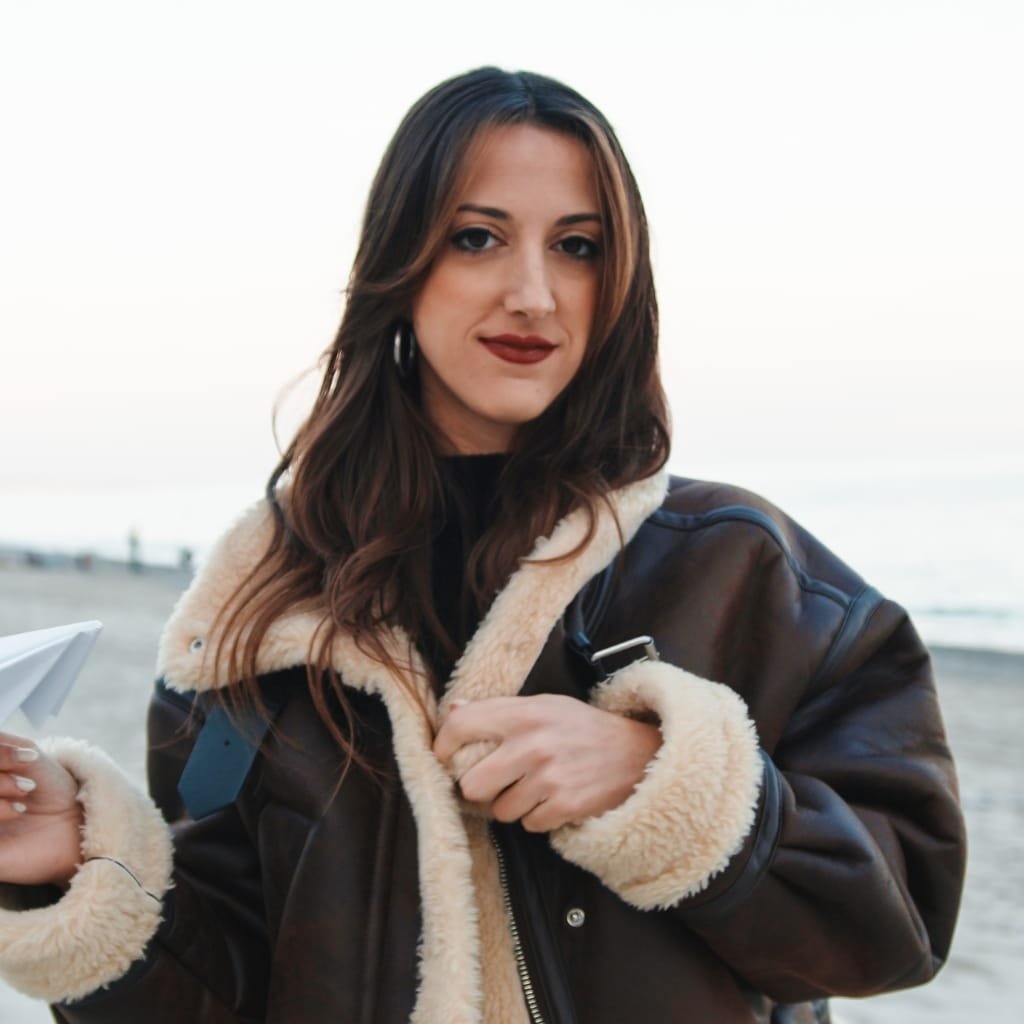 A woman standing outdoors near a beach, wearing a fashionable aviator jacket with a shearling collar, looking into the camera with a subtle smile.

