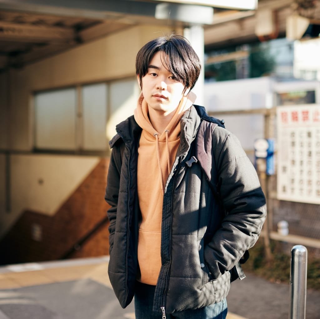 A young man standing outdoors in cold weather, wearing a black reversible bomber jacket over a peach-colored hoodie with his hands in his pockets.