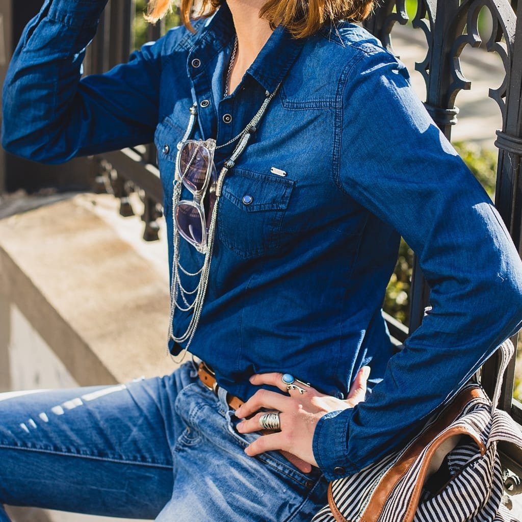 Close-up of a person wearing a blue western-inspired shirt jacket, accessorized with layered necklaces, sunglasses hanging from the collar, rings, and a striped bag, sitting outdoors with a decorative black metal fence in the background.