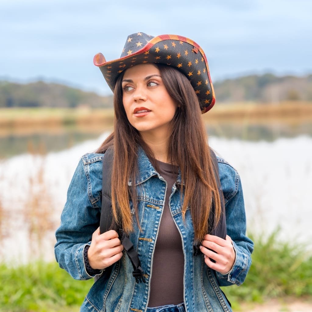 A young woman wearing a denim (western) jacket and a cowboy hat with star patterns, standing outdoors near a body of water with a blurred natural background.