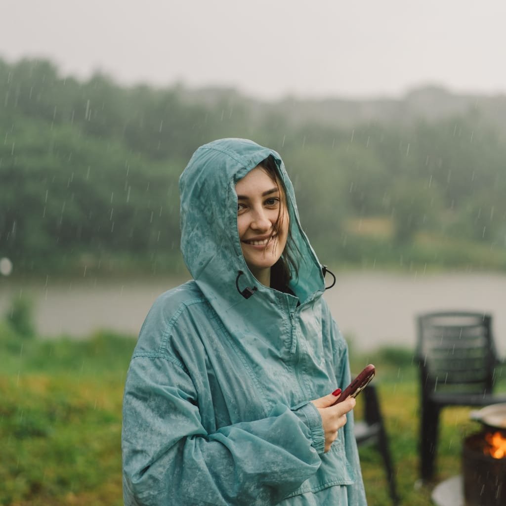 A woman wearing a teal rain jacket with the hood up, smiling while holding a smartphone outdoors in the rain near a body of water with blurred trees and outdoor furniture in the background.

