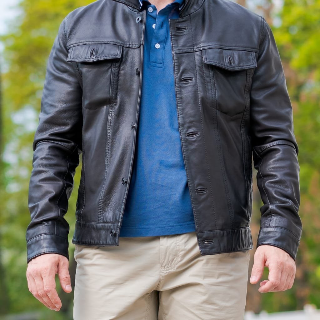 Close-up photo of a person wearing a black leather shirt jacket over a blue polo shirt and beige pants, in an outdoor setting with blurred green foliage in the background.