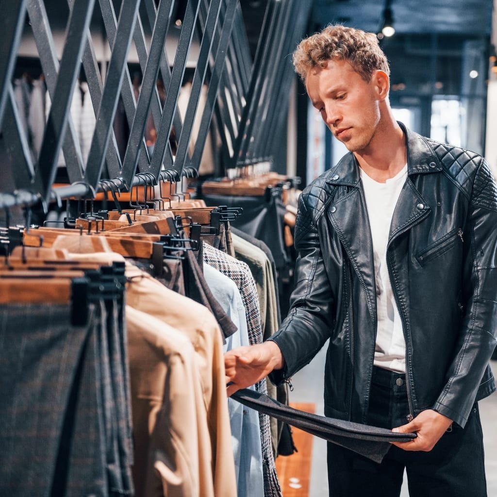 A man shopping for clothing in a store, holding a pair of pants while examining shirt jackets on a rack.