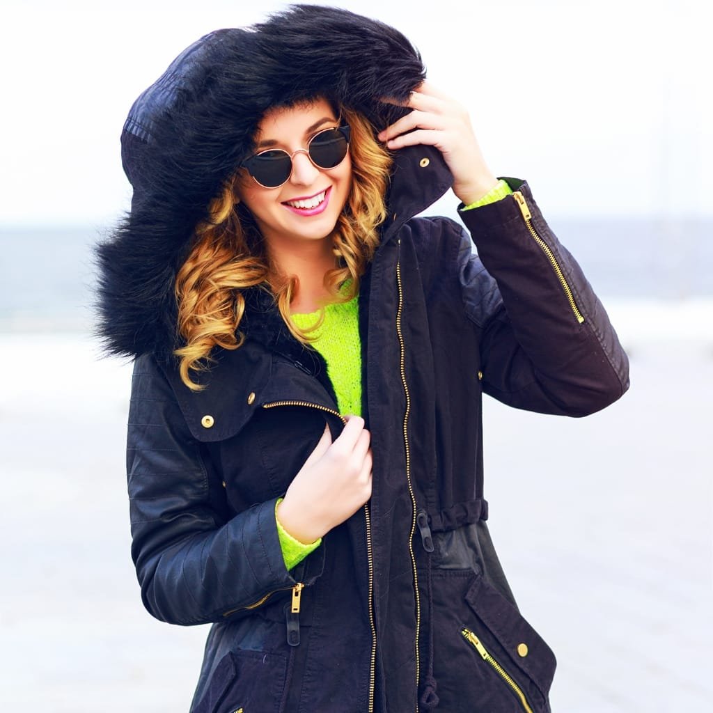 A smiling woman dressed in a black winter parka with a fur-lined hood, wearing round sunglasses, and a bright neon green sweater underneath. She is outdoors, possibly at the beach or a similar open area, adjusting her hood.

