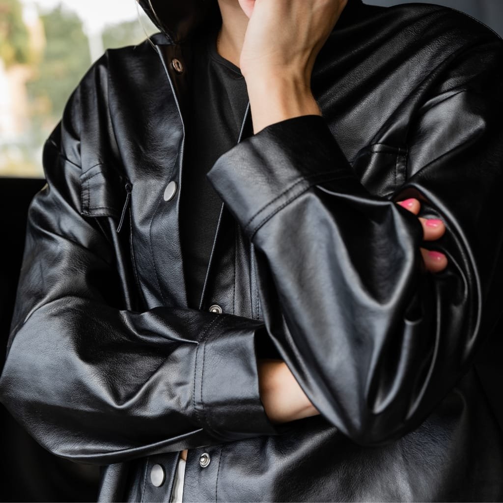 A person wearing a black leather shirt jacket, with one arm crossed and the other hand resting near their face.

