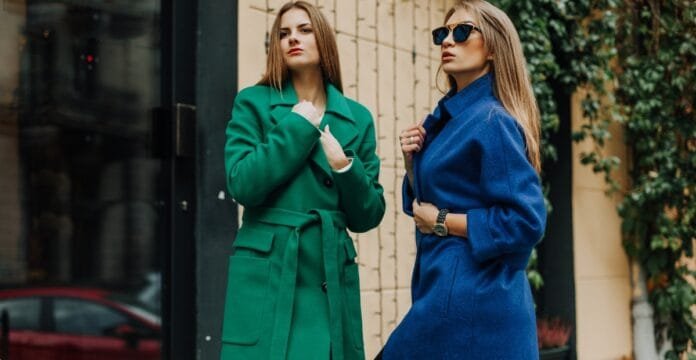 Two women are standing outdoors, wearing stylish coats. One woman is in a green belted coat, and the other is in a blue coat, both with confident poses.