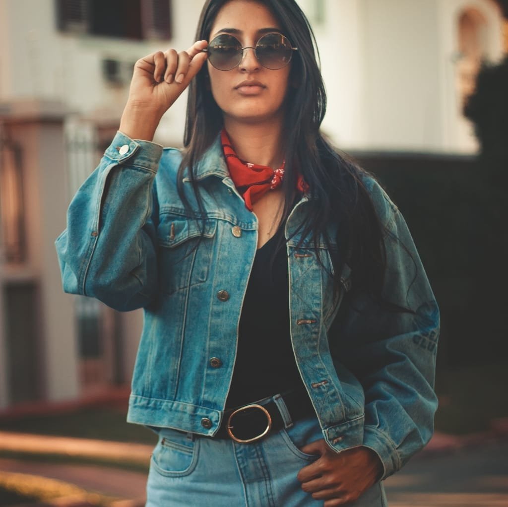 A woman with dark hair and sunglasses is wearing a classic denim jacket over a black top, accessorized with a red bandana around her neck and a belt with a circular buckle. She is standing outdoors, confidently posing with one hand on her hip and the other adjusting her sunglasses.