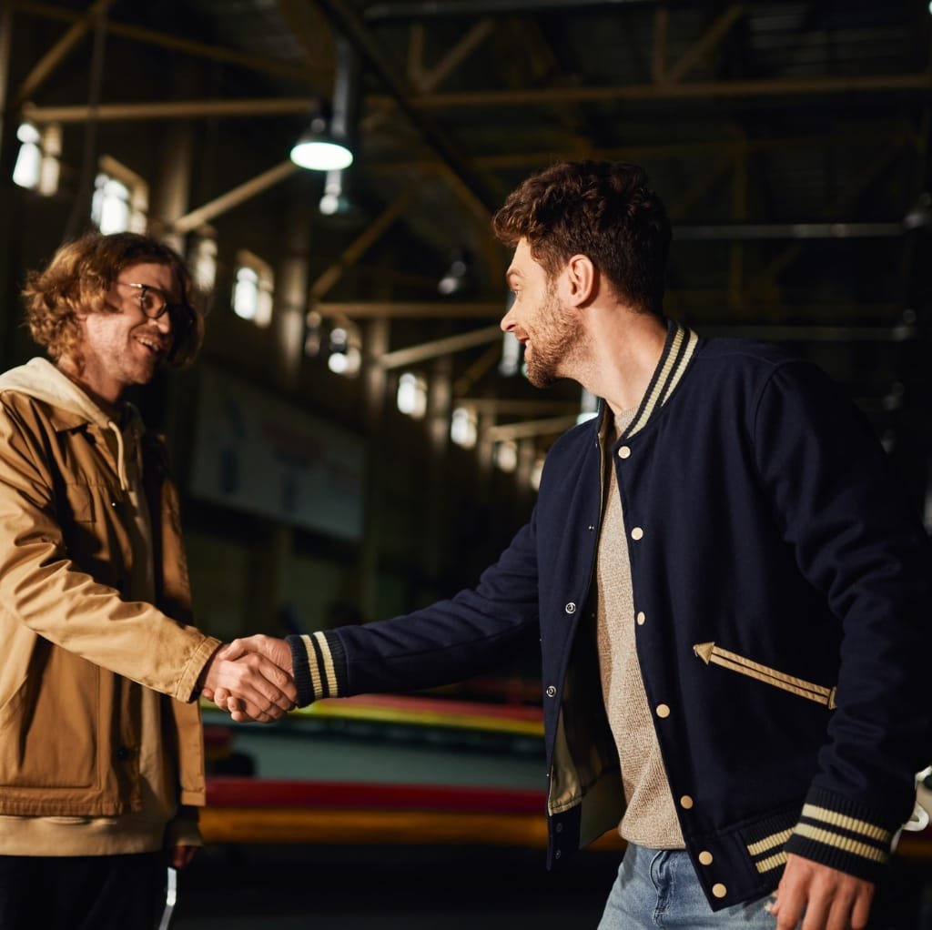 Two men shaking hands inside a dimly lit indoor space, with one wearing a navy wool varsity jacket with beige accents and the other in a tan jacket and glasses.