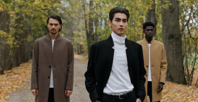 Three men wearing stylish wool work coats, walking in a wooded autumn setting.