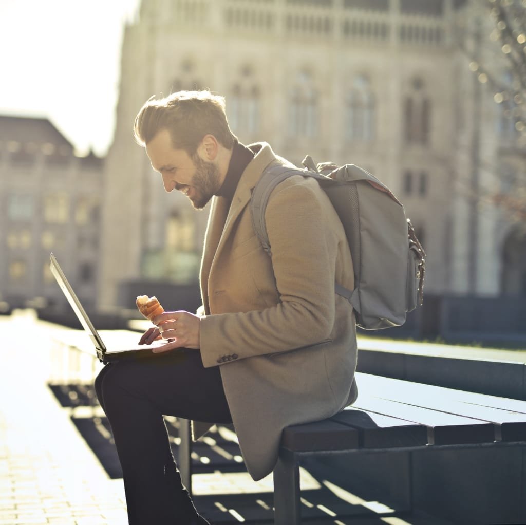 A stylish man sitting on a park bench, wearing a wool work coat and carrying a backpack, holding a pastry and looking at a laptop.