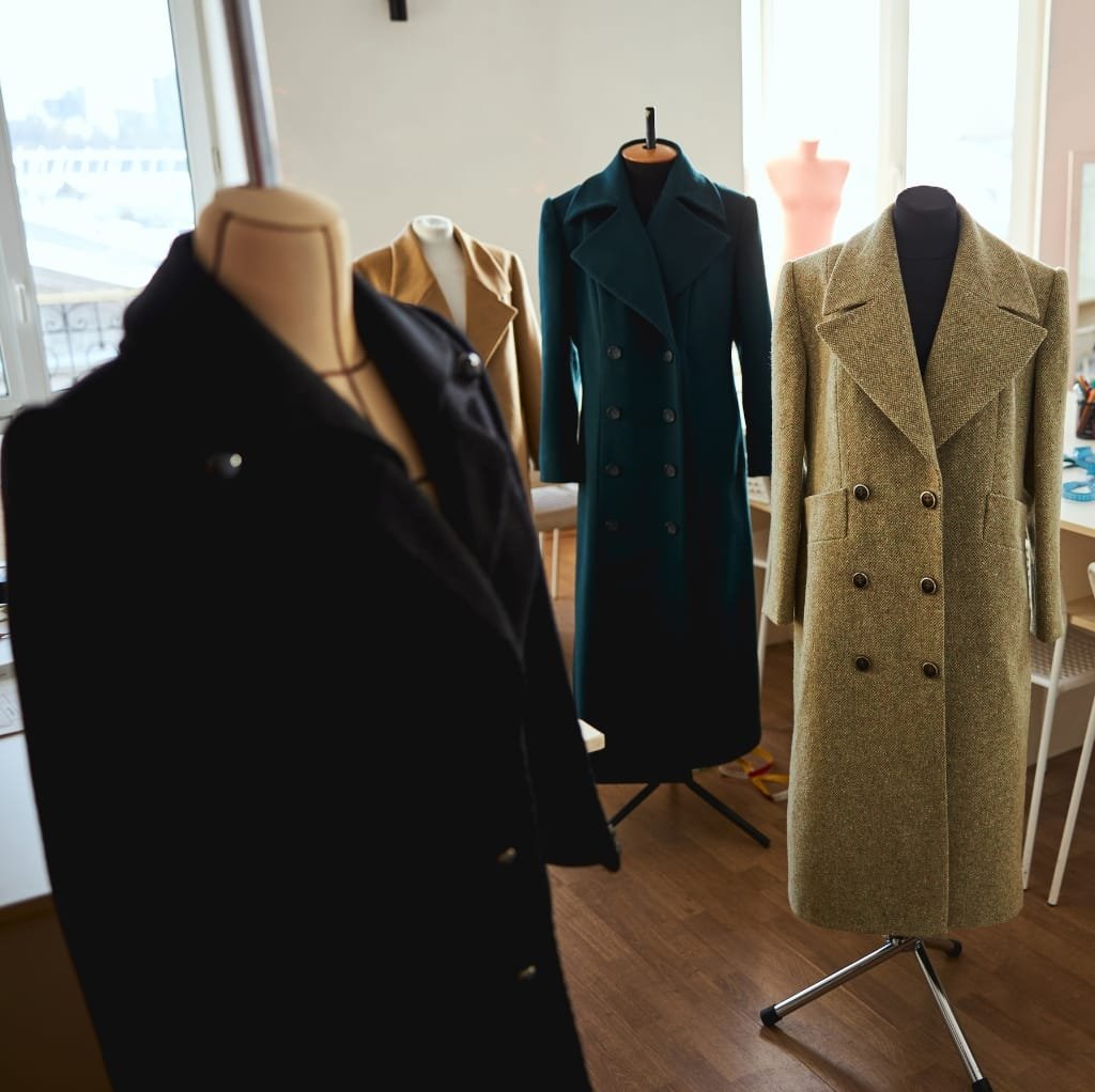 A display of wool work coats for men on mannequins, featuring various styles in colors such as black, beige, teal, and a patterned greenish-brown, set in a well-lit indoor space with large windows.
