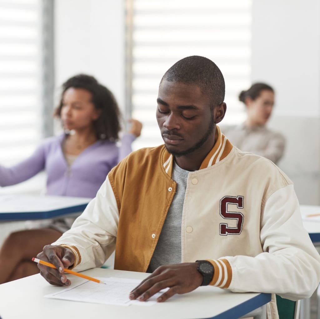 A young man in a light and dark beige wool varsity jacket with an "S" emblem on the chest, sitting at a desk in a classroom or office setting, focusing on his notes with other people blurred in the background.