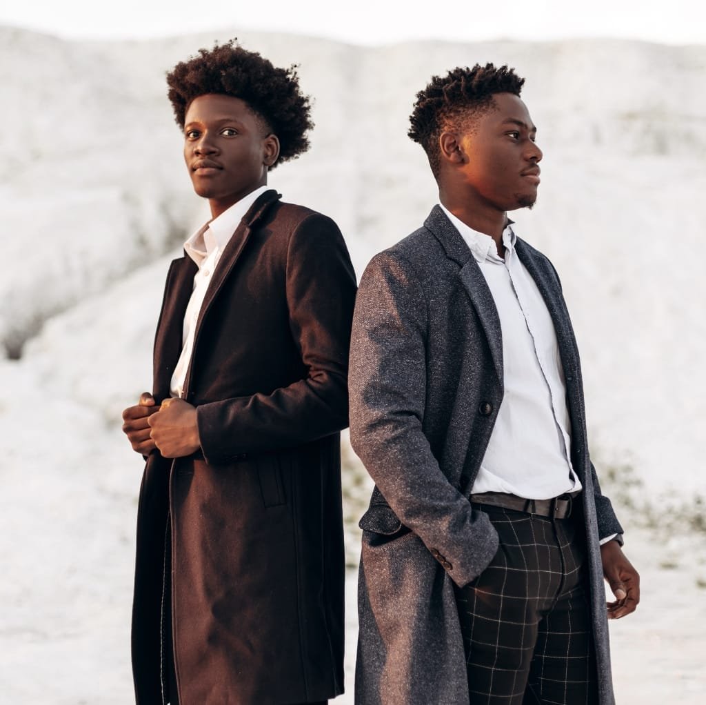 Two men standing outdoors in stylish wool coats. The man on the left wears a long, dark wool coat over a white shirt, while the man on the right sports a gray patterned wool coat with a white shirt and checked trousers.