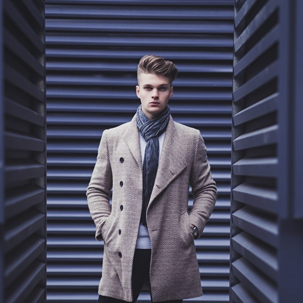 A young man with styled hair wearing a beige herringbone-patterned coat over a shirt and scarf, standing between dark, horizontal slat walls.