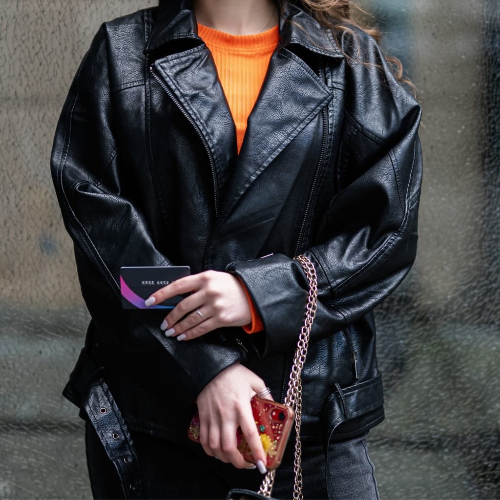 A person dressed in a black leather jacket holding a credit card in one hand and a smartphone with a decorative red case in the other, standing against a textured wall.

