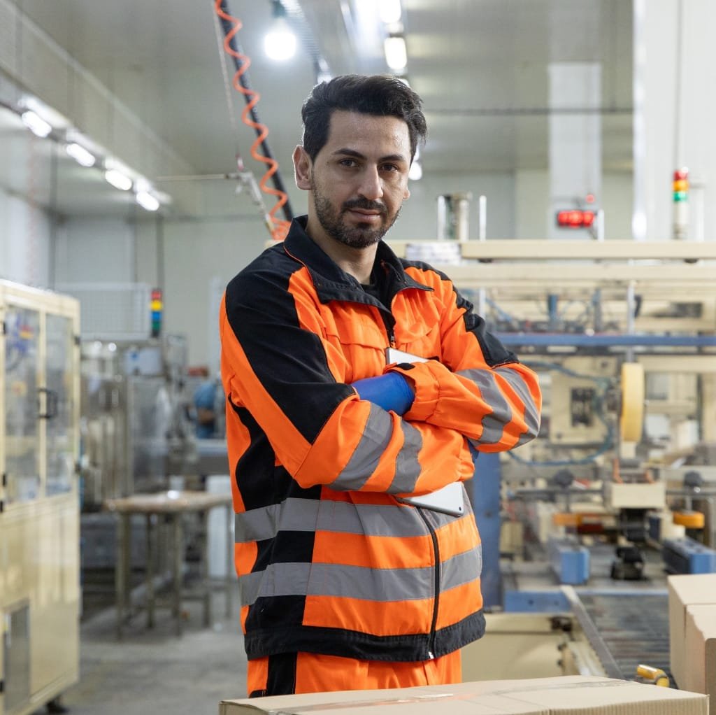 A man in an industrial setting wearing a high-visibility orange and black workwear lined jacket with reflective stripes, confidently standing with arms crossed.