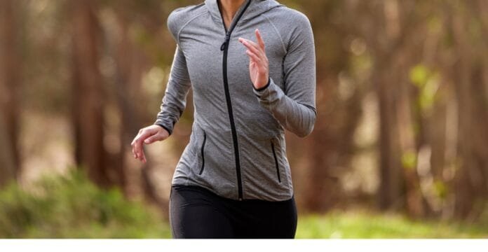 A woman wearing a grey zip-up running jacket and black leggings, jogging outdoors in a wooded area.