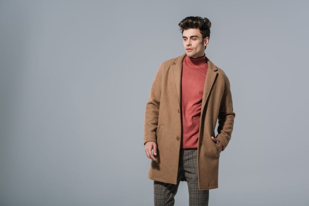 A young man wearing a stylish men's wool work coat in a light brown color, paired with a rust-colored turtleneck and plaid trousers, standing against a plain gray background.