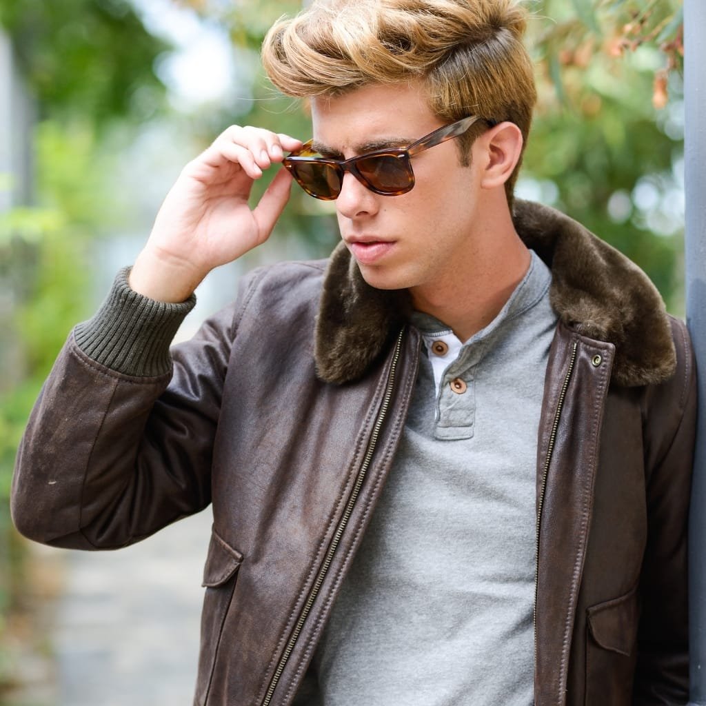 A young man with light brown hair wearing sunglasses, a gray polo shirt, and a brown leather flight jacket with a fur collar, standing outdoors.