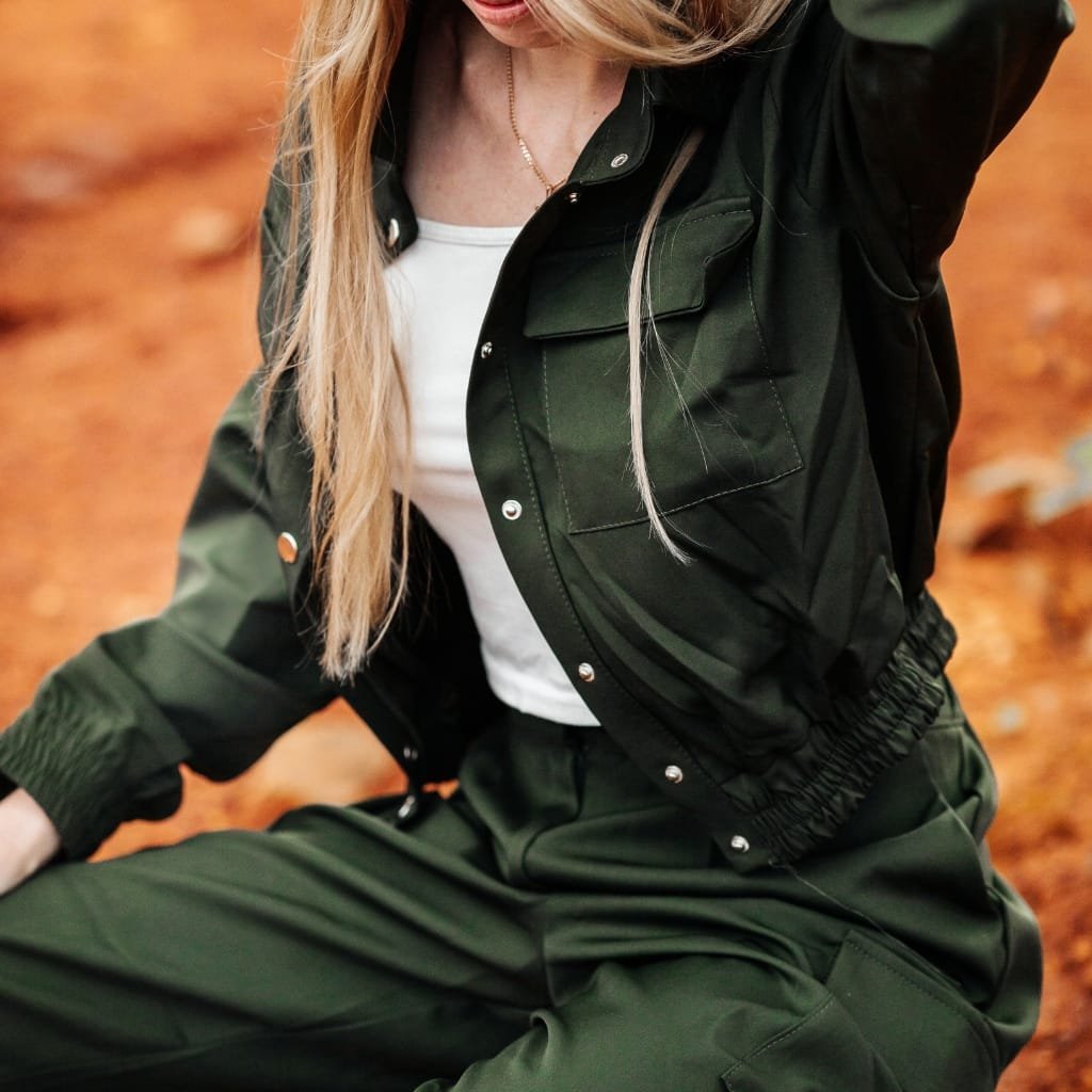 Close-up of a woman wearing a dark green flight jacket and matching pants, sitting outdoors on a fall leaf-covered ground.