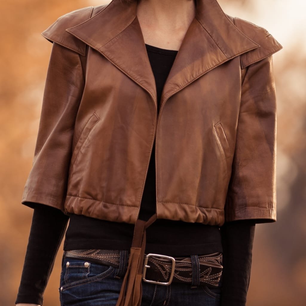 A close-up of a person wearing a stylish brown leather jacket over a black top, paired with dark jeans and a belt, set against a blurred autumnal background.