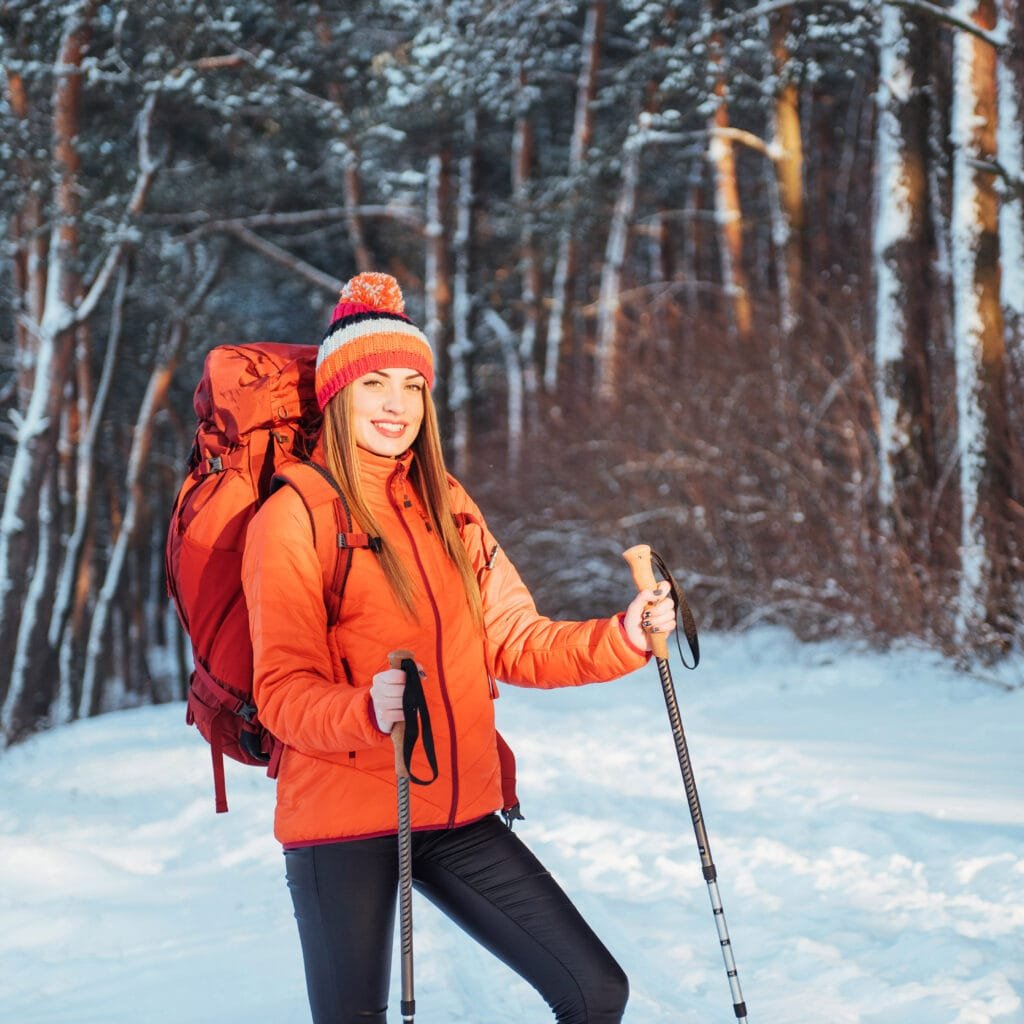 Woman wearing an Alaska winter coat hiking through a snowy forest with trekking poles and a backpack.