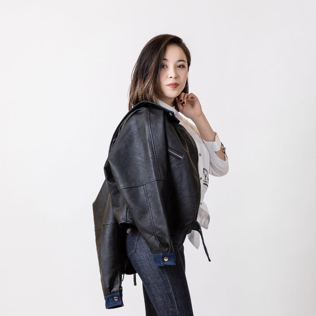 A woman wearing a black leather bomber jacket over a white shirt, paired with dark jeans, standing against a white background with a confident pose.

