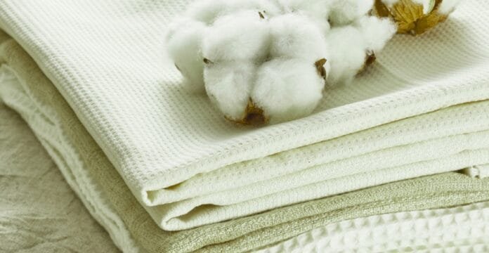 A close-up of neatly folded white and cream cotton fabrics stacked on top of each other, with cotton bolls resting on the top fabric.