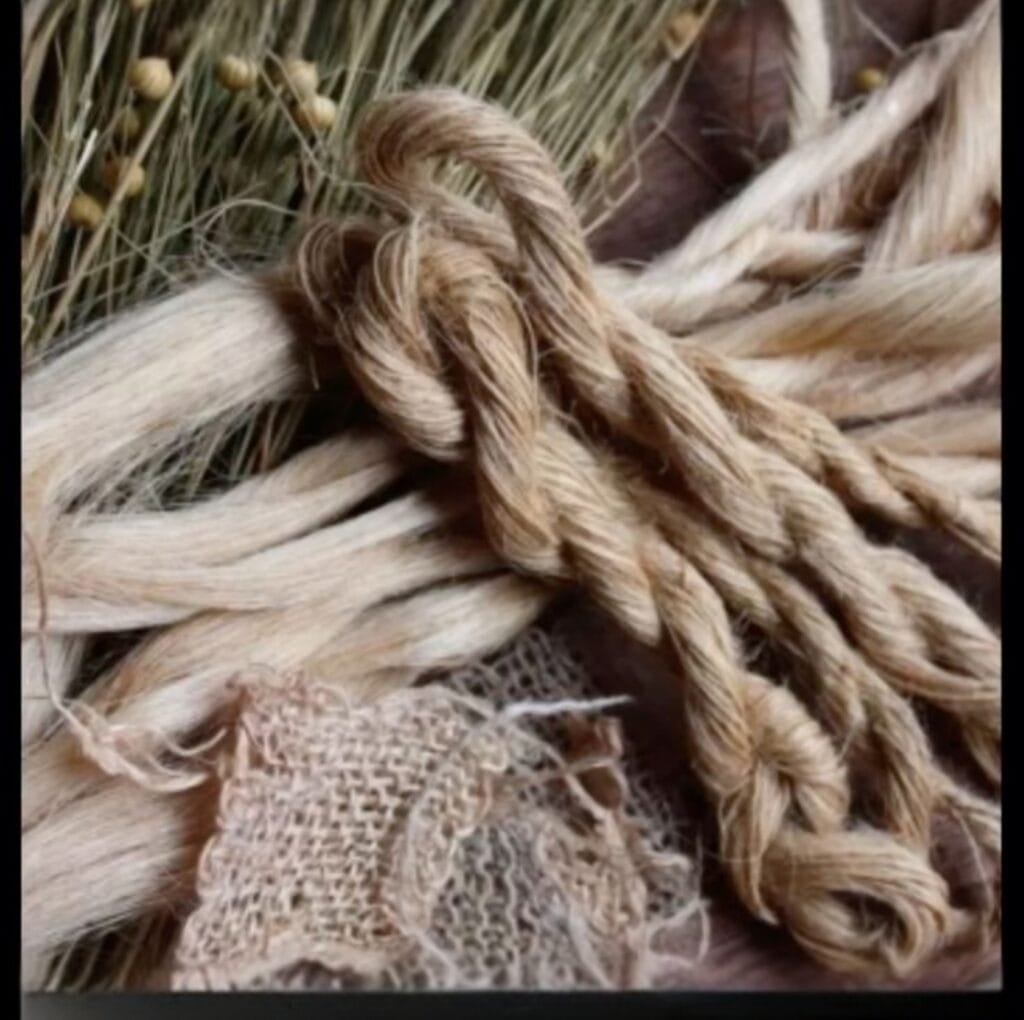 A close-up photograph of thick, natural flax fibers and twisted rope, showing the coarse beige strands and a piece of coarse woven fabric. The fibers look rustic and unfinished, with a few frayed ends.