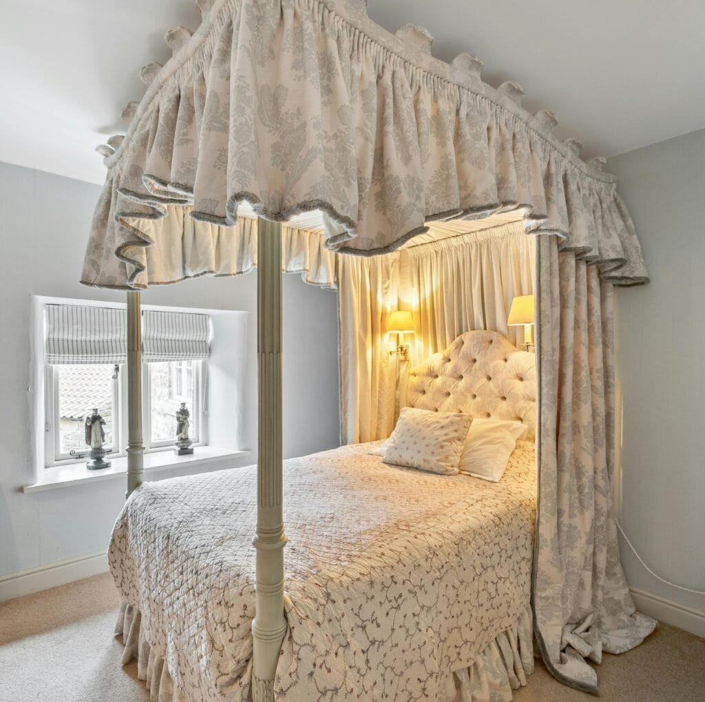 A luxurious four-poster bed with a tufted headboard, draped in cream damask fabric. The canopy features gathered edges and decorative fringe, matching the bedspread and curtains for an opulent, elegant look.