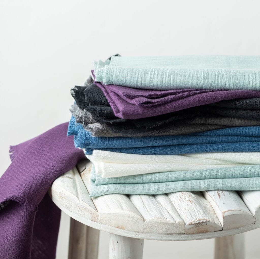 A stack of folded Belgian linen fabrics in soft, earthy tones (pale mint, lavender, charcoal, blue, and cream) arranged on a rustic white wooden stool. A single purple linen drape spills over the edge.