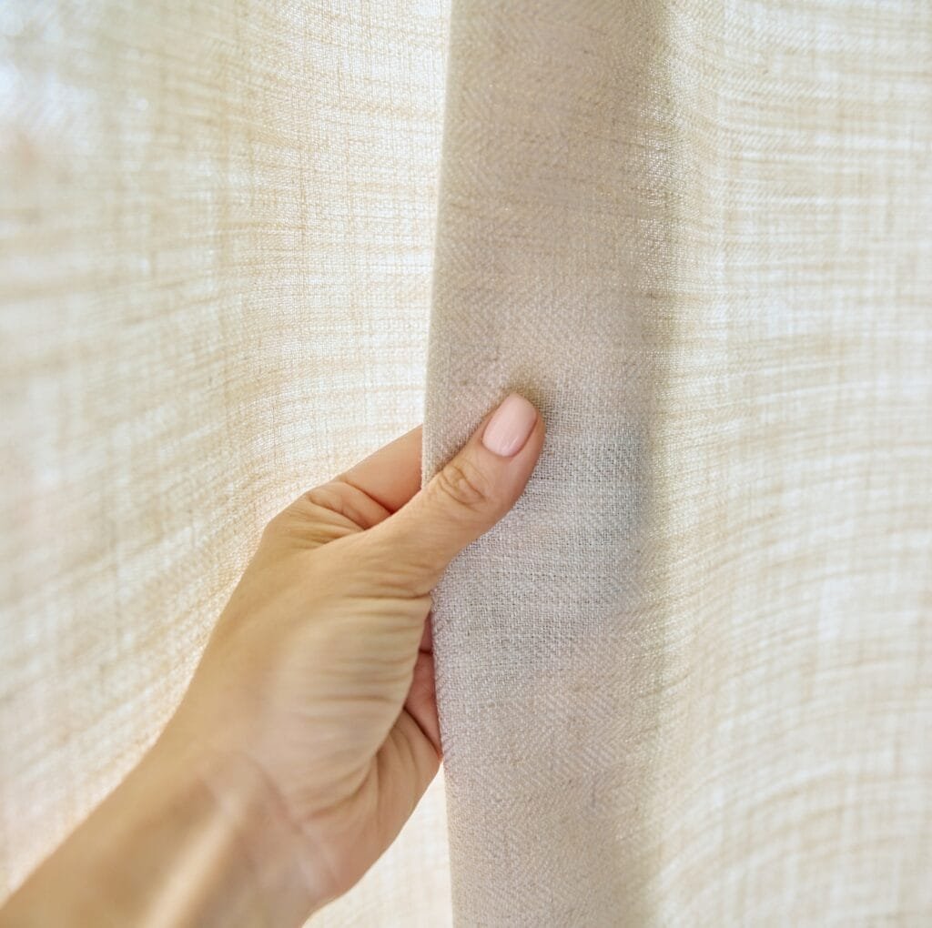Close-up of a hand holding a light beige linen curtain, showing the plain weave texture.