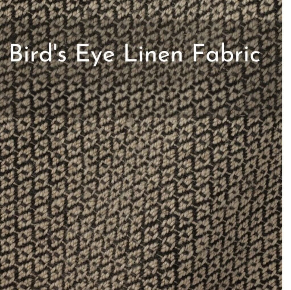 Close-up view of a textured fabric with a small, irregular geometric pattern in black and beige. The pattern creates a speckled, “bird’s eye” effect across the surface.