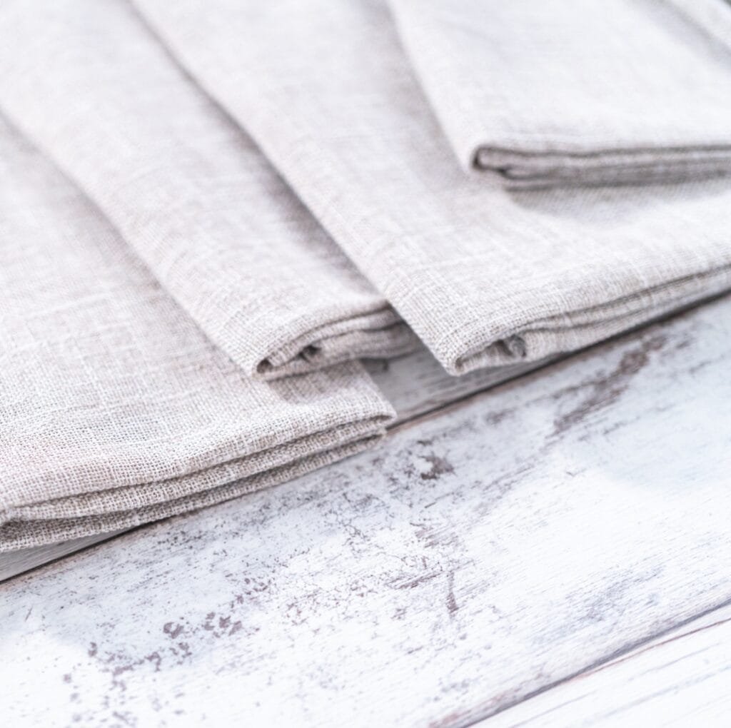 A close-up view of light beige linen fabric folded neatly on a weathered white wooden surface, showcasing the fabric’s coarse weave and natural texture.
