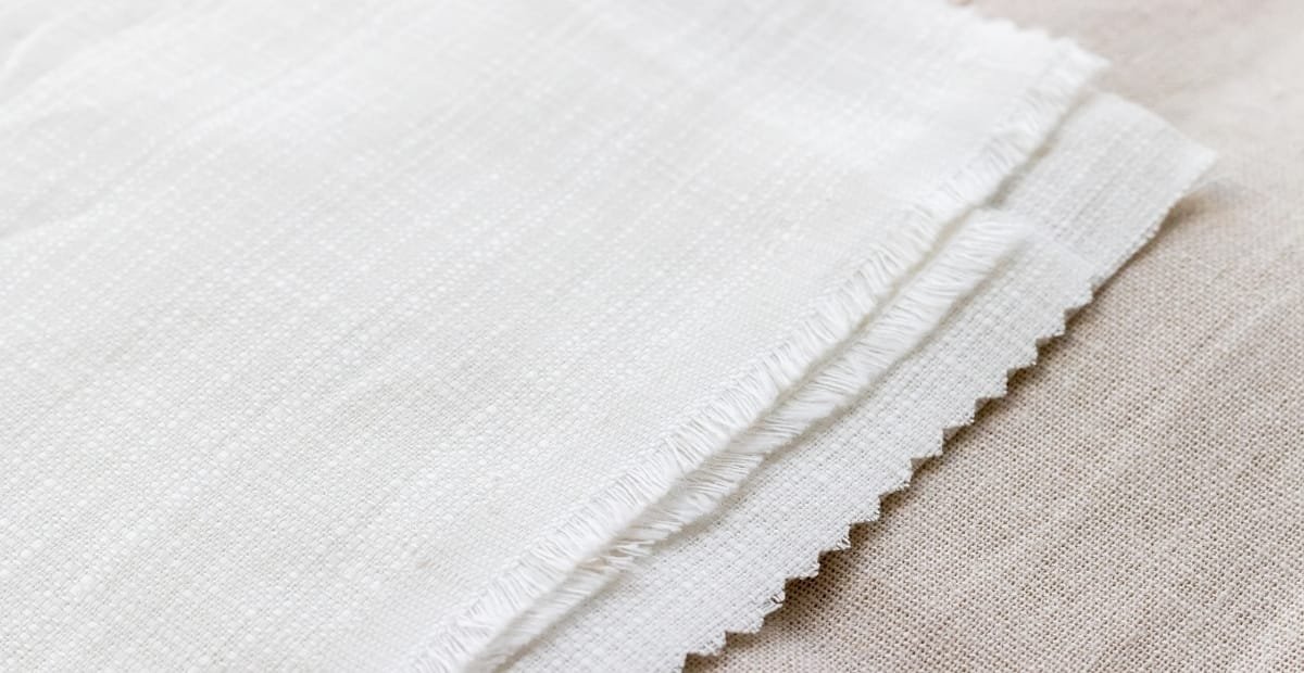 Cambric Linen fabric: Effortless Style & Enduring Durability A close-up view of white cambric linen fabric with a finely woven, smooth texture and a fringe along the edge.