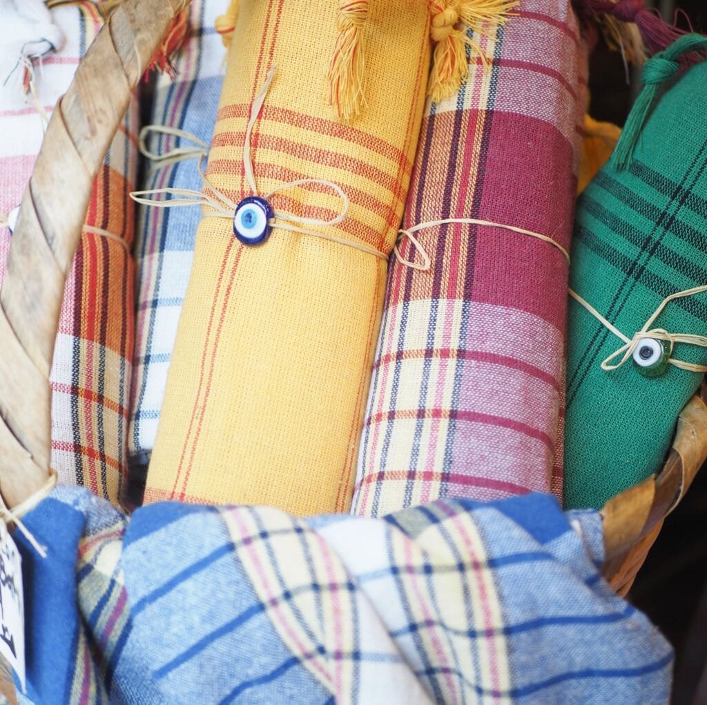 Close-up of rolled linen fabrics in bright colors (yellow, orange, red, green) tied with natural twine. Each roll has a small blue-eyed safety bead accent, resembling an evil eye, and a woven plaid pattern in varying hues.