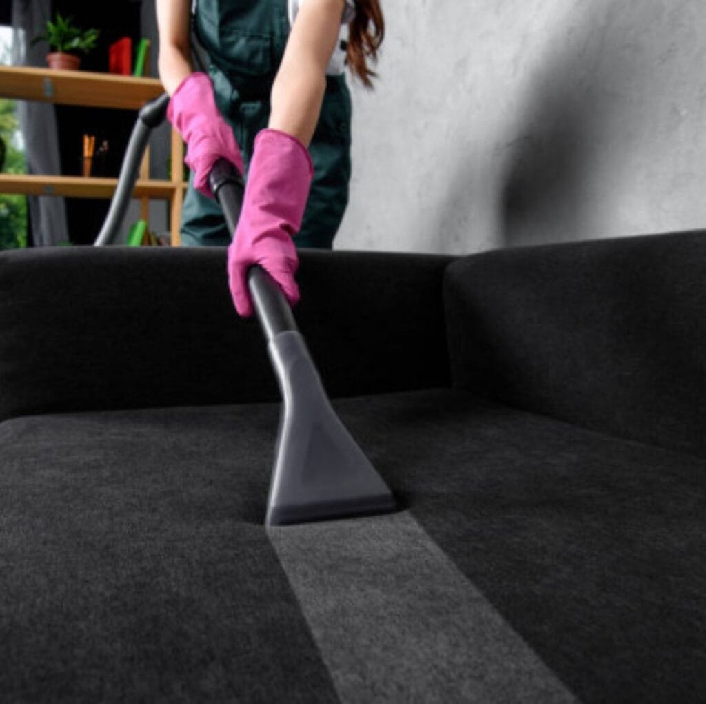 A person wearing pink gloves uses a handheld upholstery tool to vacuum clean a dark gray fabric sofa in a living room.