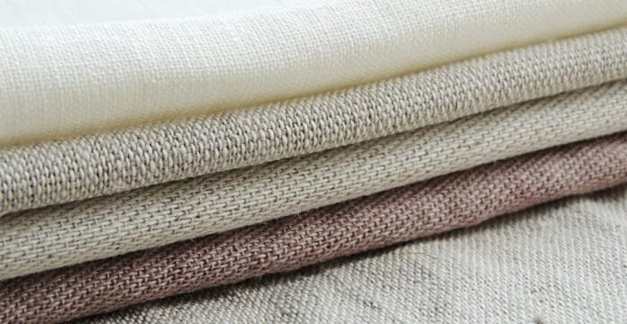 Close-up of several folded linen fabric pieces with a tight weave in neutral tones (ivory, beige, taupe, and light brown).”