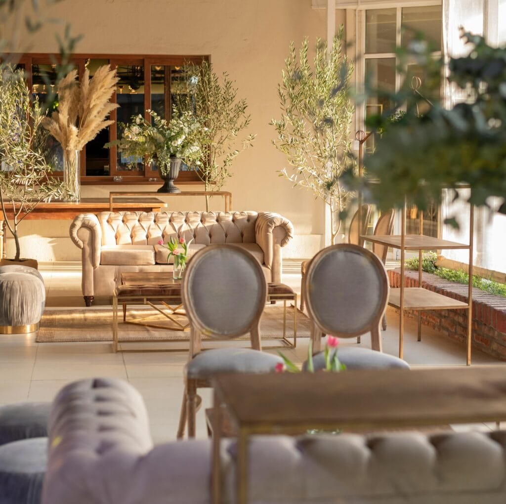 A sunlit living room with elegant, neutral-toned furniture including a tufted beige sofa, round-backed gray chairs, and a wooden coffee table. Lush greenery and tall plants frame the space, creating a warm, inviting ambiance.