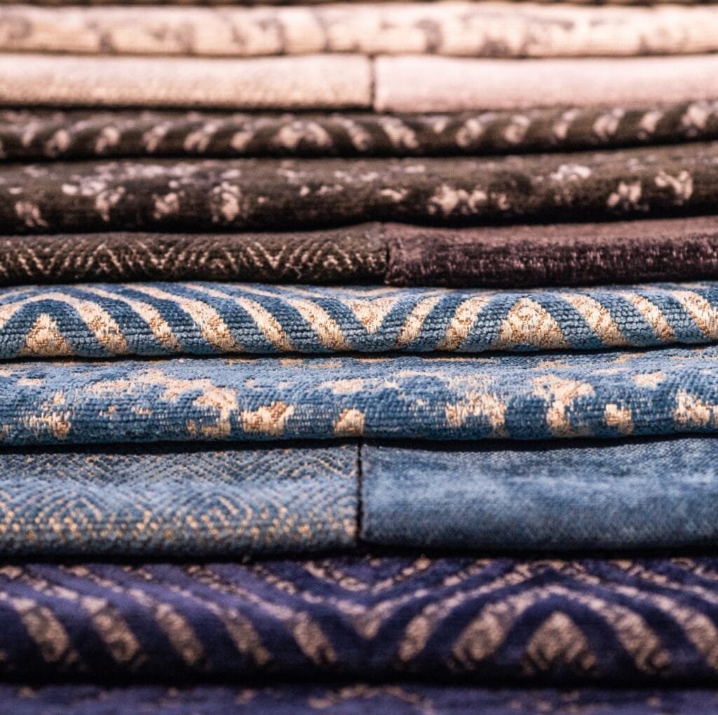 A close-up image of neatly folded damask linen fabrics in various shades of blue and neutral tones, displaying intricate woven patterns and textures.
