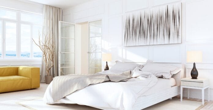 A bright, modern bedroom with a white bed, beige and white bedding, a white nightstand with a dark lamp, a yellow sofa in the corner, and large windows letting in natural light.