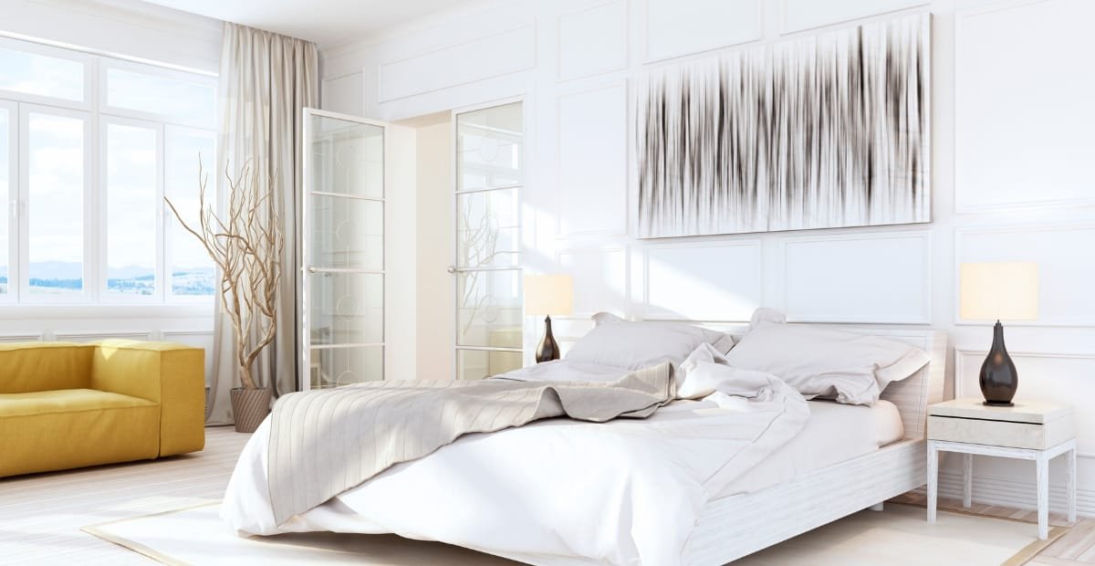 A bright, modern bedroom with a white bed, beige and white bedding, a white nightstand with a dark lamp, a yellow sofa in the corner, and large windows letting in natural light.