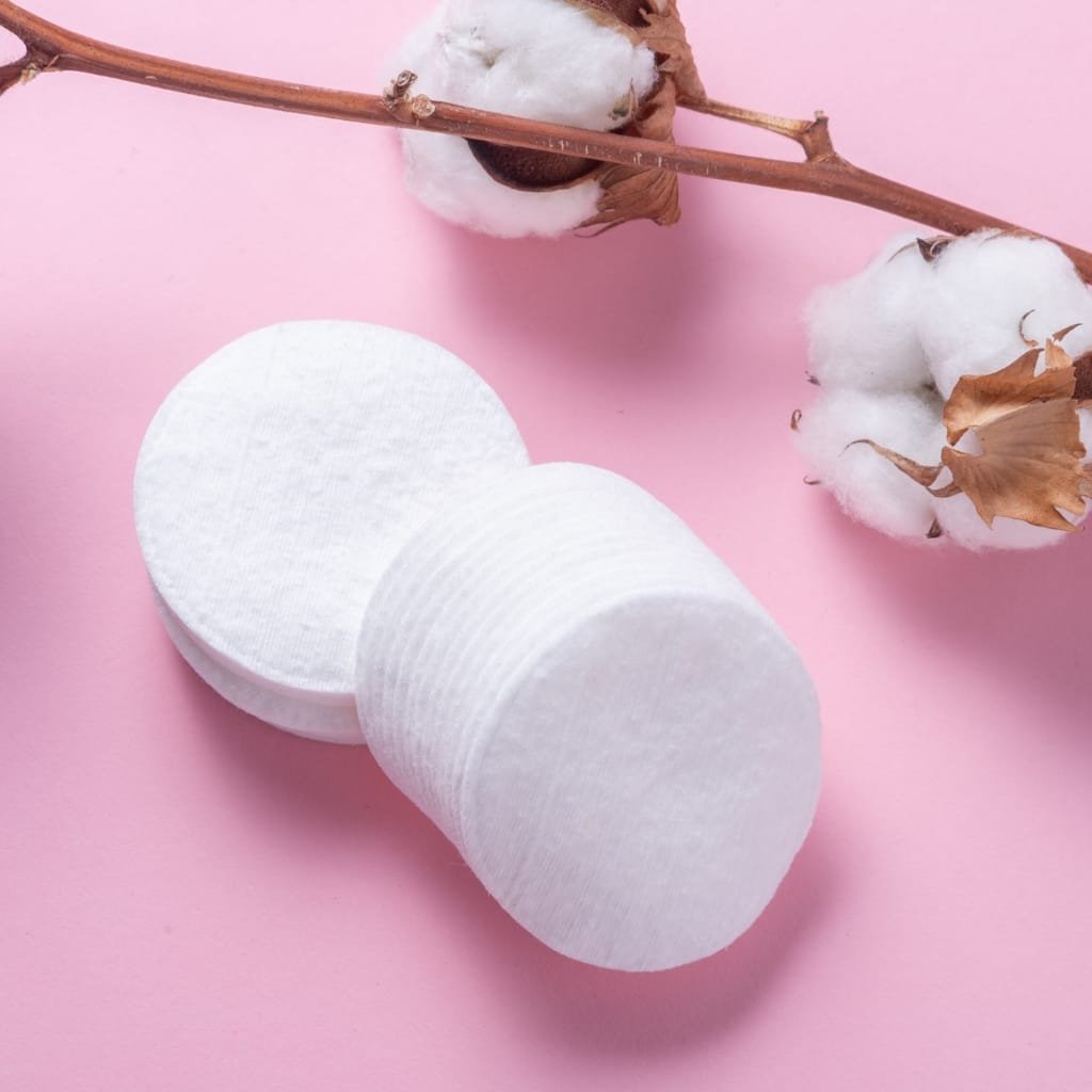 A neat stack of white cotton pads on a soft pink background, with a cotton branch featuring fluffy bolls in the top right corner.