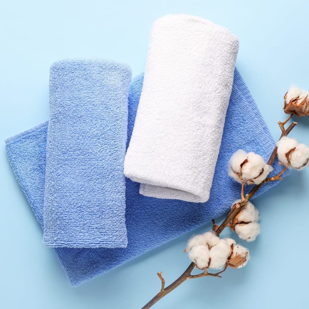 A stack of soft blue towels with a white rolled towel on top, arranged on a light blue background next to a cotton branch with fluffy bolls.

