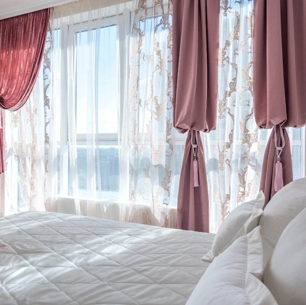 A sunlit bedroom featuring large windows dressed with layered drapes and sheer curtains. The outer curtains are dusty rose with tassels, pulled back in places, while ornate sheer panels with damask embroidery filter the light, revealing a view of a cityscape beyond. A neatly made white quilted bed with fluffy pillows sits in the foreground.