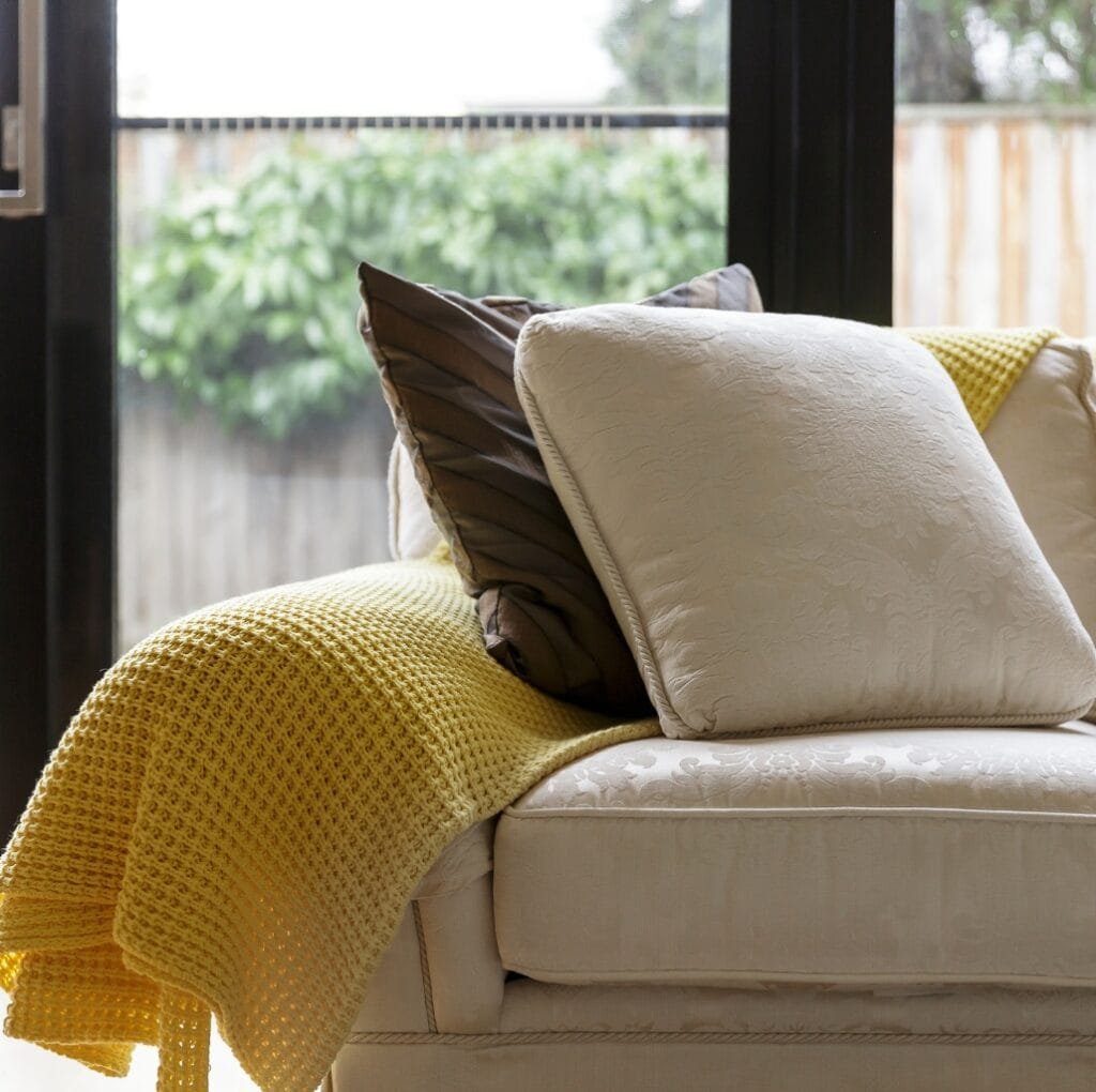 A cozy living room scene featuring a light beige upholstered sofa with decorative cushions and a bright yellow knit throw draped over the armrest, set near a large window that brings in natural light.