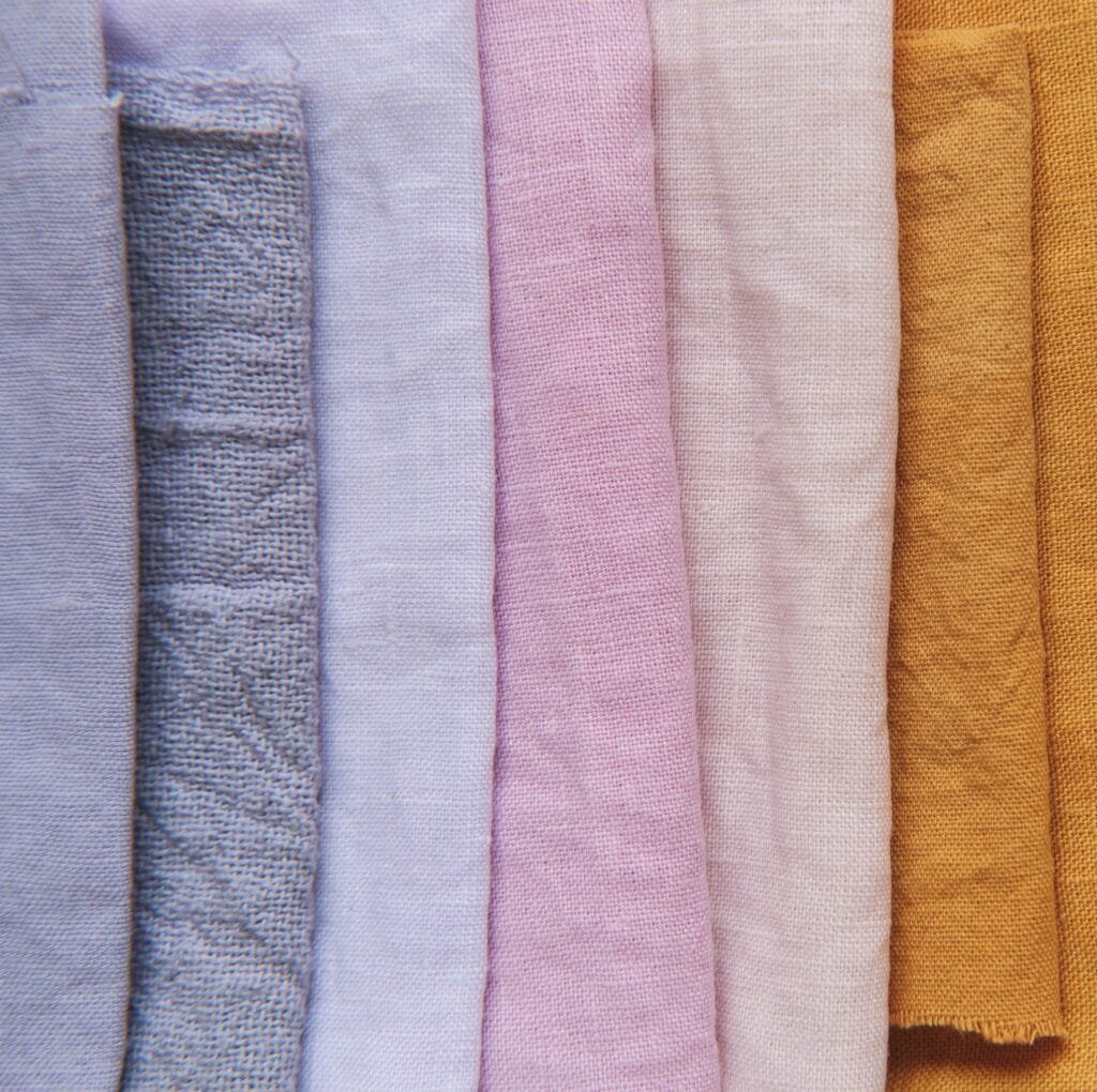 A vertical assortment of linen fabric swatches in soft pastel and warm earth tones, including light blue, powder blue, lavender, pink, cream, and golden tan, showcasing the natural texture of linen.