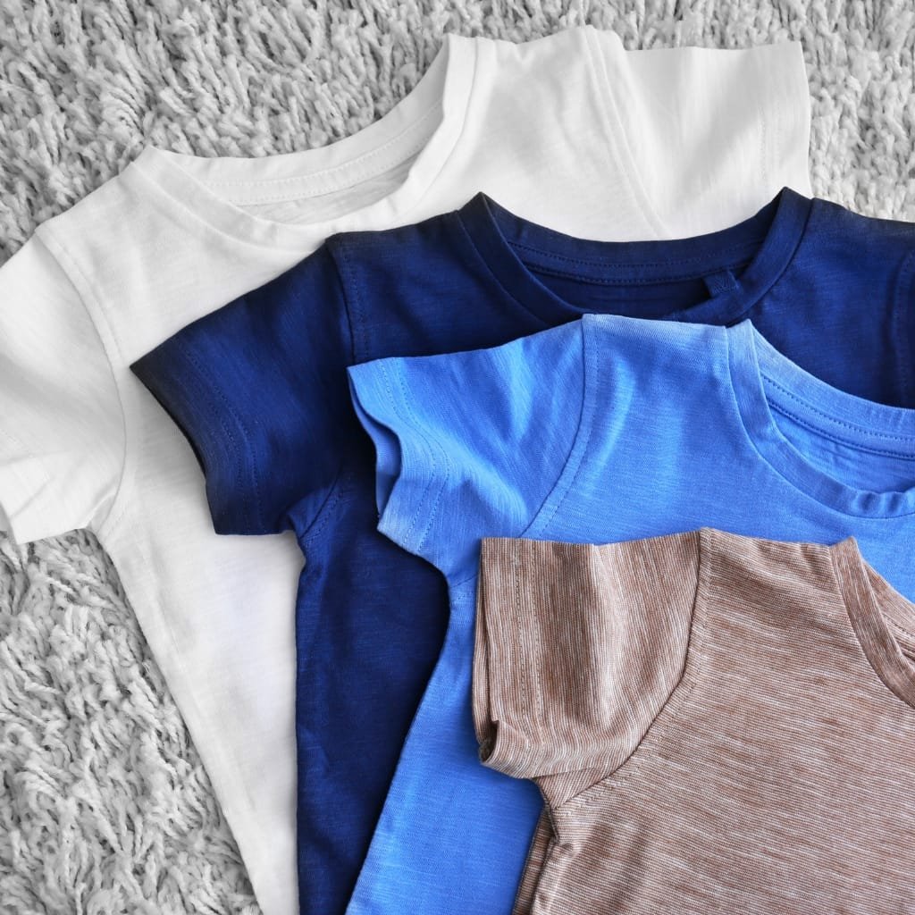 A stack of neatly folded cotton T-shirts in white, navy, blue, and heather gray/brown tones spread on a soft, textured gray rug.