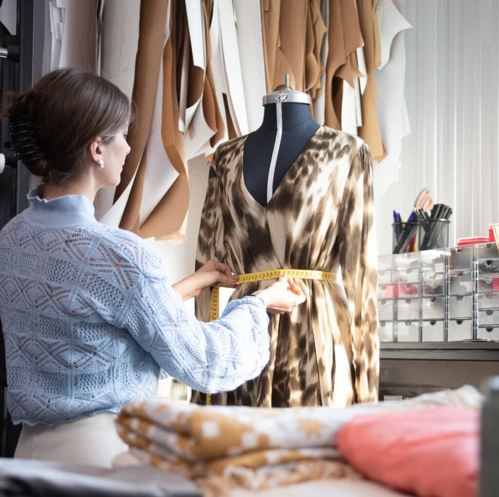 A fashion designer or seamstress measures a flowing, animal-print linen dress on a dress form in a bright studio. Linen fabric scraps hang in the background, and a yellow measuring tape is wrapped around the waist area.