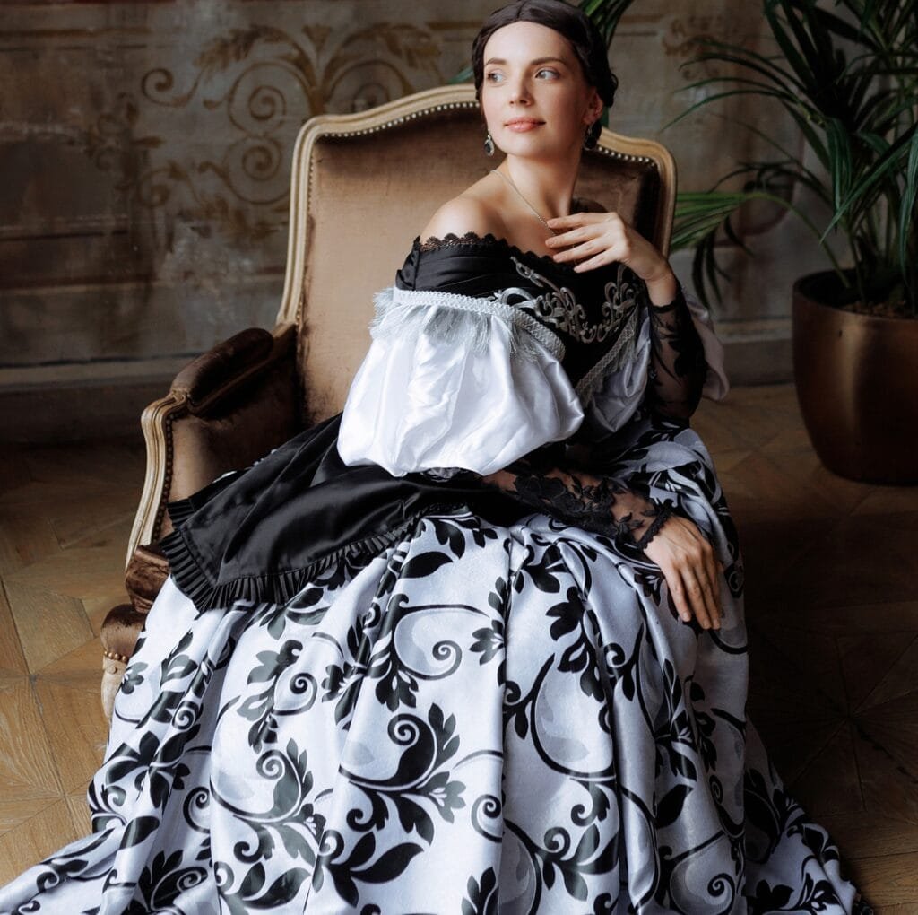 A woman sits gracefully in an ornate vintage chair, wearing a dramatic black-and-white damask linen gown with puffed white sleeves and delicate lace details. The patterned skirt flows across the wooden floor, creating a luxurious, historical look.